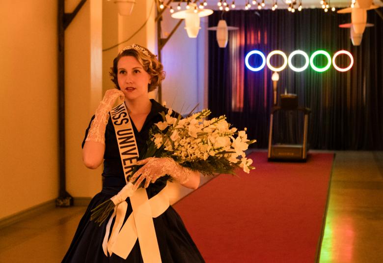 Kuvassa on käytävässä kävelemässä sinisessä mekossa kukkakimppu kädessään ja valkoinen nauha, jossa lukee Miss Universum.  Käytävän takana on viisi kirkasta ympyrää. 