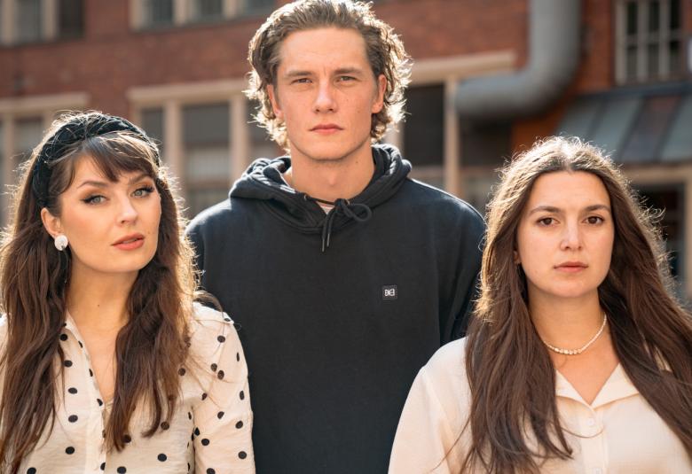 Kuvassa ovat puolivartalokuvassa Rita Behm valkoisessa sini-pallollisessa puserossa, Pyry Kähkönen mustassa hupparissa ja Mimosa Willamo vaaleassa puserossa.  Taustalla on punatiilinen kerrostalo, josta näkyy ikkunoita.