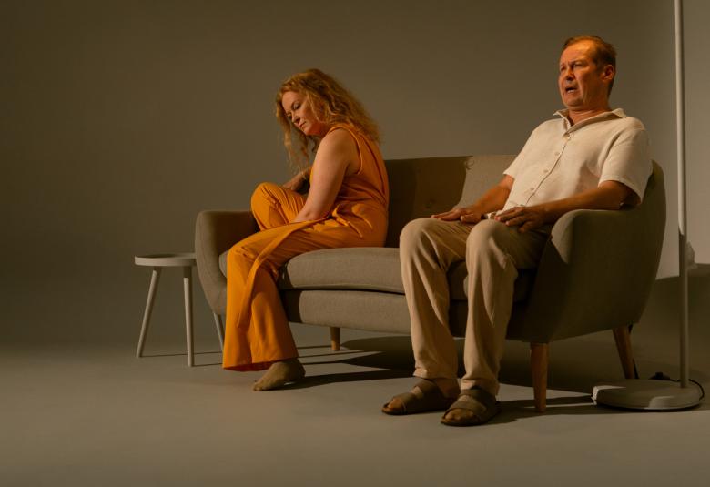 Kuvassa on Meri Nenonen istumassa harmaalla sohvalla vasemmassa nurkassa sivuttain ja kumartuneessa asennossa. Hänellä on oranssi housuhaalaripuku päällään. Oikealla istuu Niko Saarela ja hänellä on valkoinen paita sekä harmaat housut.