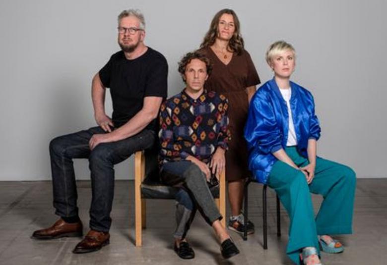 Kuvassa on vasemmalla Roland Persson istumassa tuolin käsinojalla ja hänellä on musta t-päitä ja farkut. Hänen vieressään oikealla istuu jani Ruscica musta-ruskea kuviollisessa paidassa ja mustissa farkuissa. Hanna Vihreälä seisoo takana ruskeassa mekossa ja edessä oikealla istuu tuolilla sinisessä puserossa sekä vihreissä pitkissä housuissa.