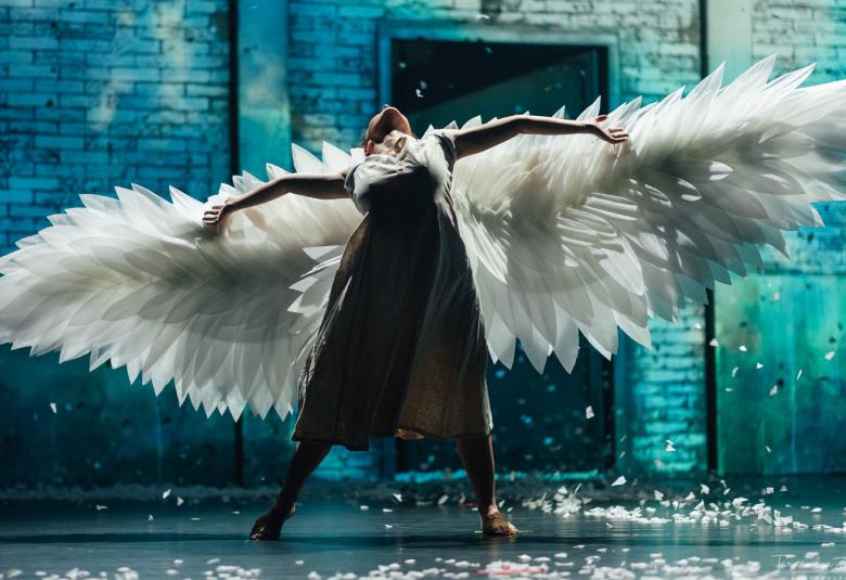 Kuvassa on mustan joutsenen esittäjä ja hänellä on isot valkoiset siivet levitettynä. Taustalla on turkoosia seinää.