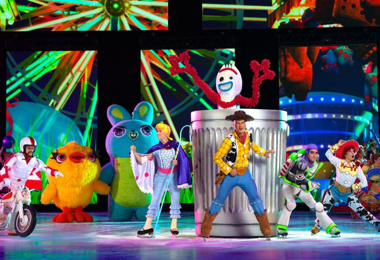 Kuvassa on etualalla jäätanssijoita valkoisissa puvuissa ja niiden oikealla puolella on Toy Storyn kolme hahmoa värikkäissä vaatteissa.  Taustalla on keltainen kuvio, josta tulee keltaisia säteitä. 