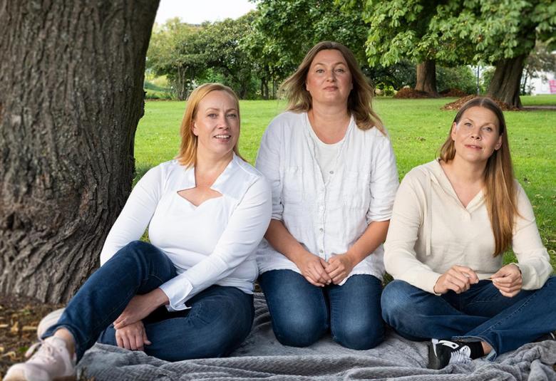 Kuvassa ovat istumassa ruohikolla puistosa suuren puunrungon vieressä peiton päällä Kaisa Hela, Ria Kataja ja Eeva Soivio.  He ovat sinisissä farkuissa ja valkoisissa paidoissa. 