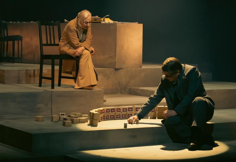 Kuvassa on vaalea, porrastettu näyttämö ja siihen osuu valoa keskelle.  Alhaalla oikealla on Timo Torikka istumassa portaalla ja kokoamassa kaarevaa pienoismallia. Ylemmällä portaalla istuu tuolilla Seela Sella kumartuneena kohti Torikkaa ja juttelee hänelle. Sellalla on vaalean ruskea puku. 