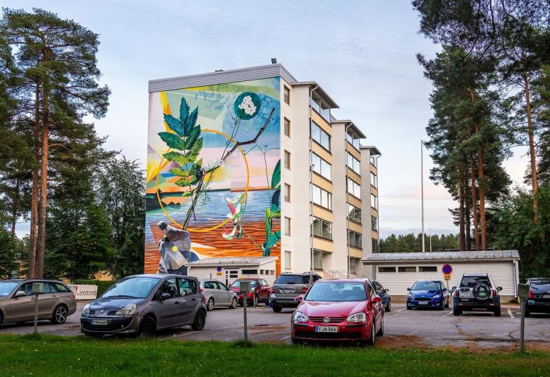 Kuvassa on kerrostalo ja sen päädyssä on taideteos, jossa on sinistä Oulujokea, vasemmalla iso vihreä pihlajanlehti ja pokealla akileija. Taistalla on vaalean sinistä ja kellertävää sekä sinitaivaalla hyppivä kauris. Pihalla on vasemmalla männikköä ja edessä autoja parkkipaikalla.