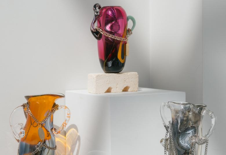 Kuvassa on kolme hyllyllä olevaa lasimaljakkoa, joista ylin on punainen, vasemmalla oranssi ja oikealla alhaalla sinertävä maljakko.  Kaikissa on kierteisiä muotoja ja ketjuja.