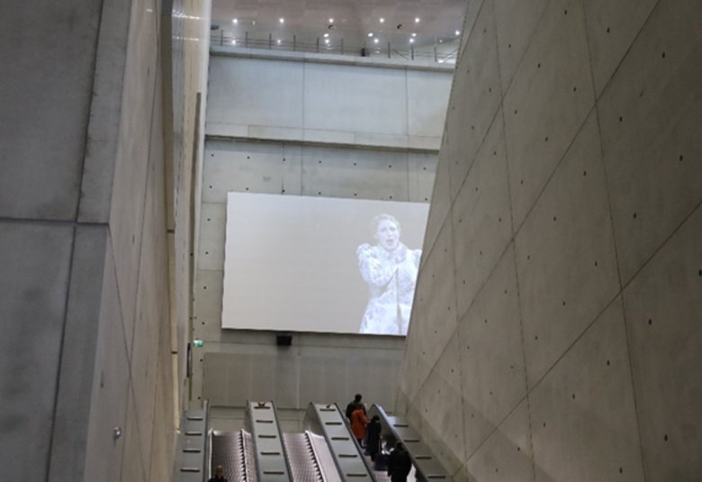 Kuvassa on Liukuportaat harmaabetonisessa käytävässä.  Yläpuolella on iso näyttö, jossa näkyy oopperalaulajamies vaaleassa vanhanaikaisessa takissa.