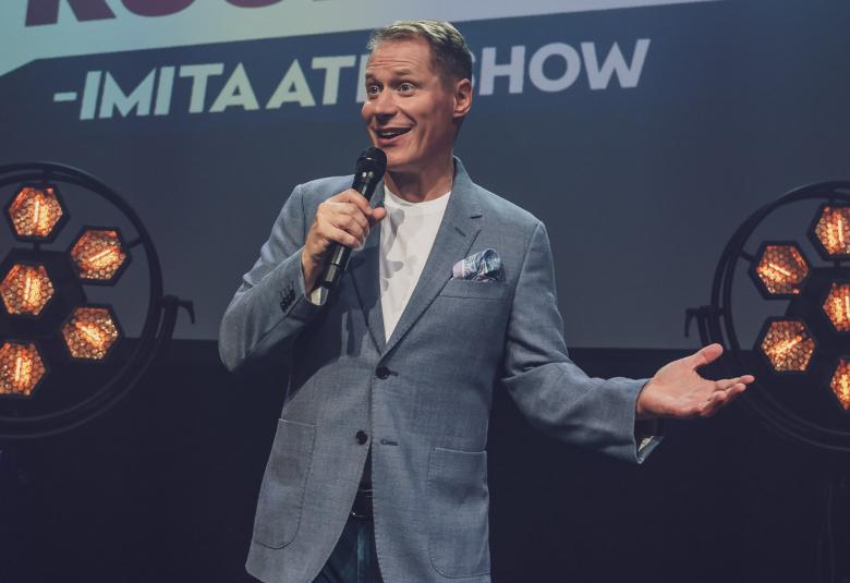 Kuvassa on Jarkko Tamminen seisomassa ja esiintymässä harmaa puku päällään ja mikrofoni oikeassa kädessään. Taustalla lukee Imitation show ja Tammisen molemmin puolin on oranssiset valokukat ympyrän sisällä sinisessä valovarressa.