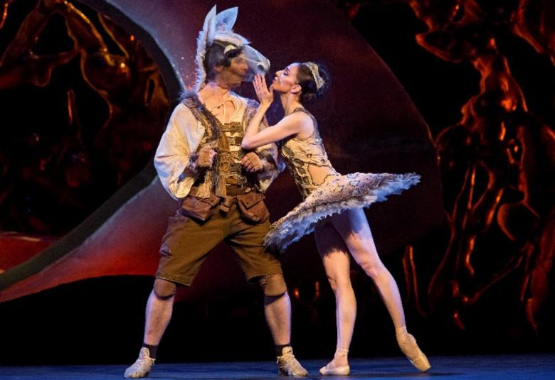 Kuvassa on tummaa, värikästä satumetsämäistä taustaa. Edessä on metsästäjäasun ruskeissa polvihousuissa ja keltaisessa paidassa sekä läpinäkyvässä hevosenpää päässään. Oikealla on sivuttain kullän värisessä puvussa naistanssija, joka pitelee sivelee hevosen turpaa.