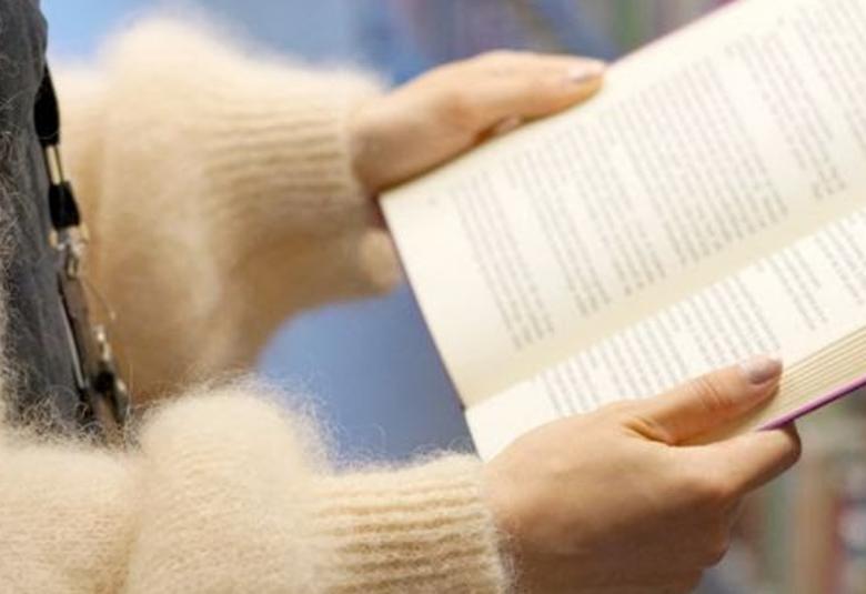 Kuvassa on naisen kädet pitelemässä avattua kirjaa. Hänellä on pörröinen, vaalea villatakki päällään. 