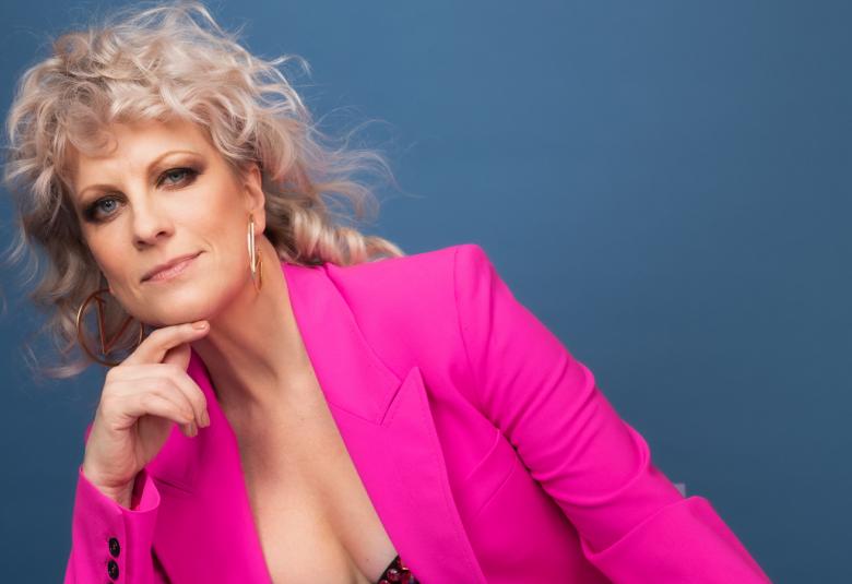 Kuvassa on Laura Vuotilainen ja hän istuu vinosti vasemmalla ja nojaa oikealla kädellään leukaansa. Hänellä on anilliini jakku. Tausta on tumman sini-harmaa.