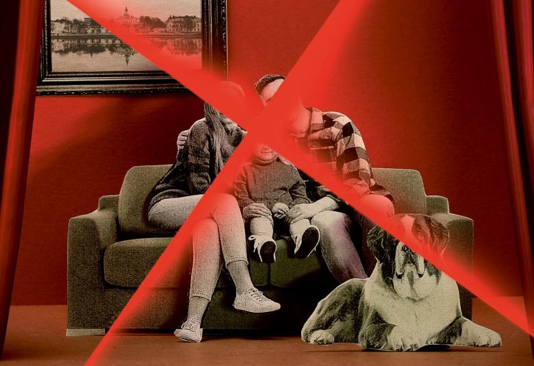 Kuvassa on nainen ja mies istuvat harmaalla sohvalla ja heidän välissään lapsi.  Seinän väri on punainen ja sillä on taulu. Pariskunnan päällä on iso punainen rasti. Oikealla lattialla on pahvikuva isosta bernhardilaisesta koirasta.