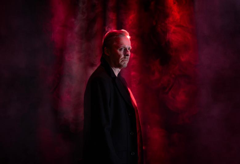 Kuvassa on keskellä tummana seisomassa puolivartalokuvassa ja valoa tulee hänen kasvoilleen sekä taustalle. Suurin osa kuvasta on tummaa.