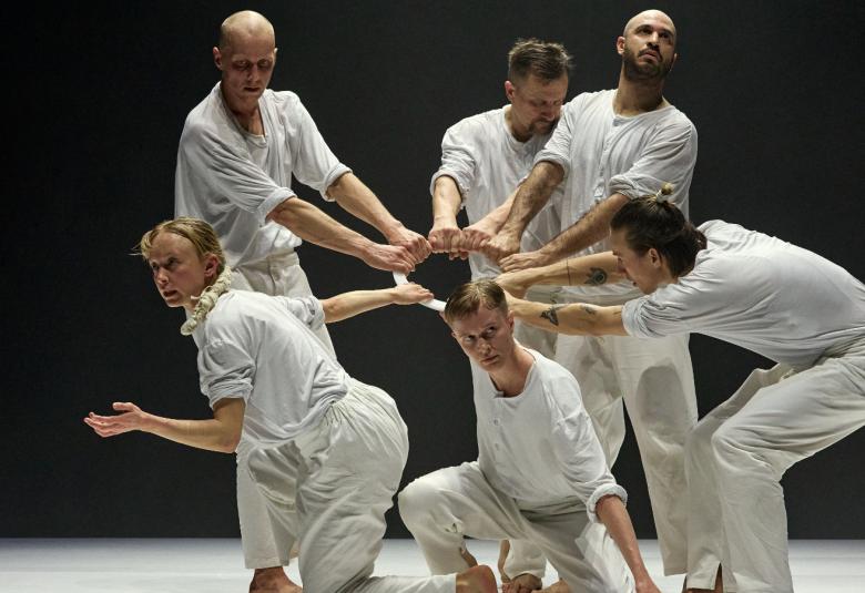 Kuvassa on kuusi tanssijaa valkoisissa asuissa ja heillä on kädet ojennettuna siten, että ne tekevät pienen ympyrän keskelle ja tanssijat ovat myös ympyrässä ja edessä olevat alempana. Tausta on tumma.