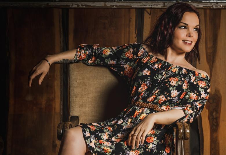 Kuvassa on Maria Tyyster ja hänellä on mustapohjainen oranssikukallinen mekko. Hän on rennosti poikittain tuolilla ja oikea käsi tuolin selkänojalla sekä katse oikealle.