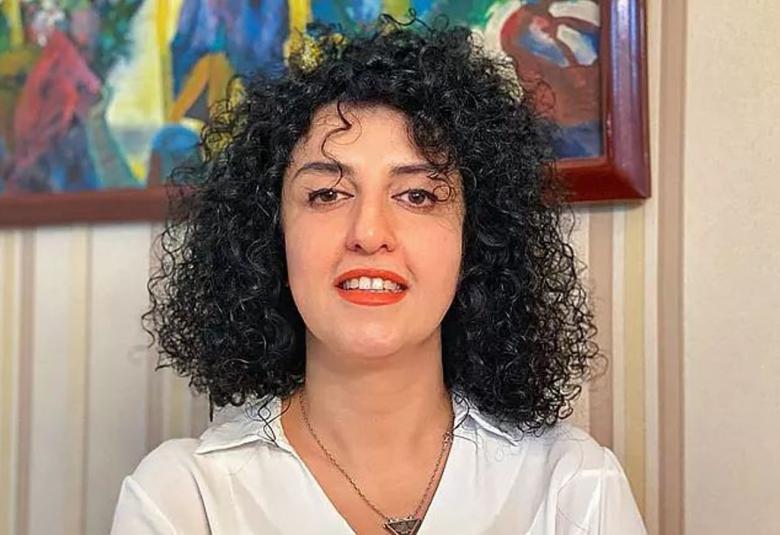 Kuvassa on Narges Mohammadin puolivartalokuva.  Hänellä on mustat kiharat hiukset ja valkoinen kauluspaita päällään. Taustalla on alaosa värikkäästä taulusta.