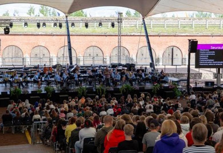 Kuvassa on lavalla poliisisoittokunta vaalean sinisissä paidoissa soittamassa. Edessä on selin yleisöä nousevassa katsomossa.  Taustalla näkyy tiilistä bastionin seinämää, jossa on ylhäältä pyöristettyjä puuovia.  Päällä on vihreää nurmikkoa. Päällä on telttakattoa.