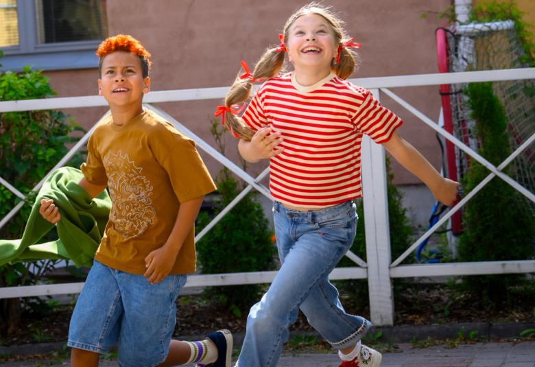 Kuvassa ovat juoksemassa kadulla Aleksi Johansson vasemmalla ja hänellä on ruskea t-paita ja farkkushortsit. Oikealla on Alma Järvensivu puna-valkoraitaisessa t-paidassa ja sinisissä farkuissa. Taustalla on valkoista ristikkoaitaa ja kerrostalon punaista tiiliseinää.