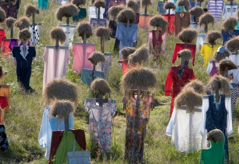 Kuvassa on vihreällä heinäpellolla ihmishahmoja, joilla on heinätupsut päänä ja vaatteet heinäseipään päällä. Hahmoja on koko kuvalalla.