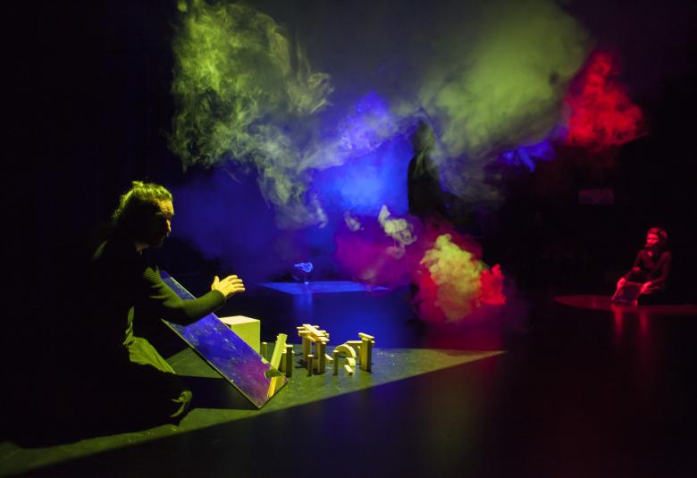 Kuvassa on tumma näyttämö ja siinä istuu lattialla mies, joka pitelee peiliä. Edessä on  keltaisessa valossa puupalikoista tehtyja portteja ja taustalla punaista savua ja ylempänä keltaista savua.  Oikealla taustalla on katselija polvillaan.