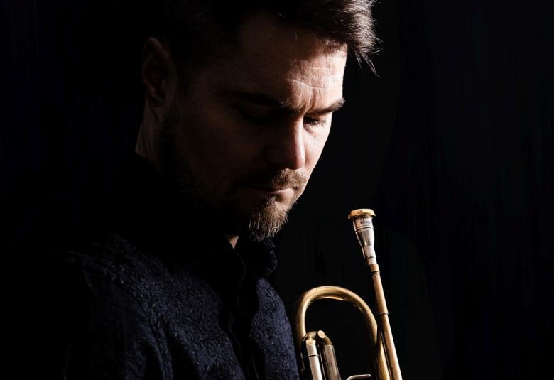 Kuvassa on Verneri Pohjola ja hänestä näkyvät kasvot sivuttain alaspäin.  Kuva on tummasti sävytetty ja kasvojen lisäksi näkyy trumpettia.