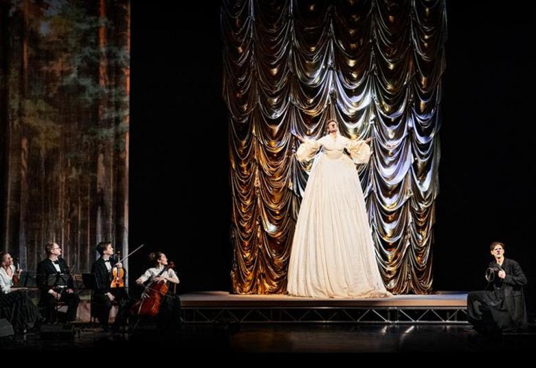 Kuvassa on näyttämöllä korkea-asuinen valkoisessa mekossa oleva mies ja vasemmalla ovat jousiorkesterin soittajat, jotka katsovat häntä. Oikealla on yksi tummissa vaatteissa oleva mies näyttämön alapuolella. Tausta on tumma.