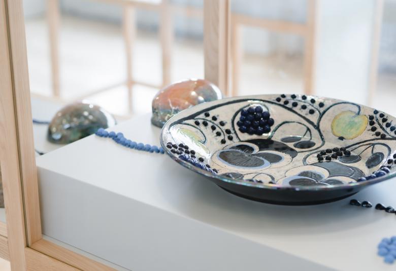 Kuvassa on lasivitriinissä Birger Kaipaisen keramiikkalautanen, jossa on valkoisella pohjalla tummansinisiä nauhoja ja rypälekuvioita ja vaalean vihreitä omenoita. Taustalla näkyy toisia vitreenejä.