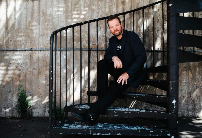 Kuvassa on Cristian Rönnbacka istumassa portailla, joissa on takana mustat pystykaiteet. Taustalla on harmaata seinää ja siihen osuu hajavaloa.  