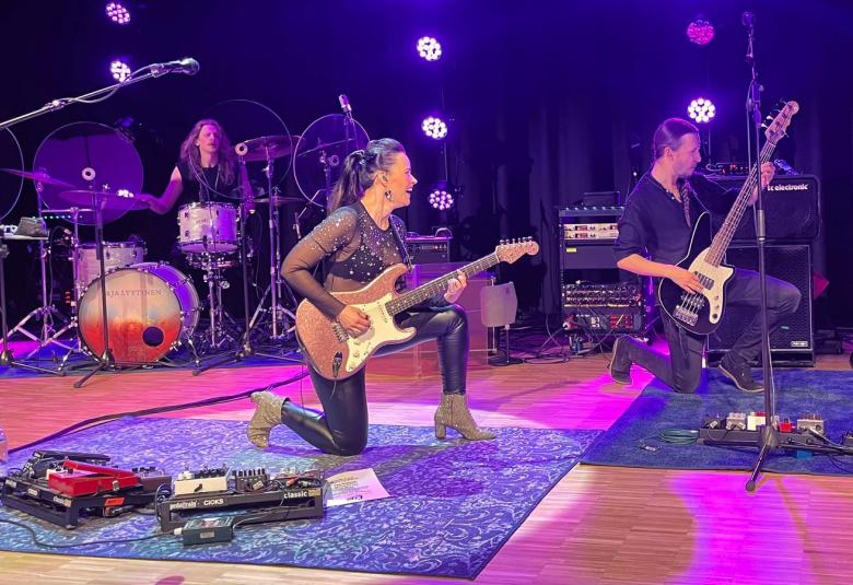 Kuvassa on Erja Lyytinen soittamassa vasen polvi lattiassa beigen väristä kitaraa. Vieressä on basisti samassa asennossa. Lattialla on sininen matto. Kuva on otettu sivusta. Taustalla on rumpali.