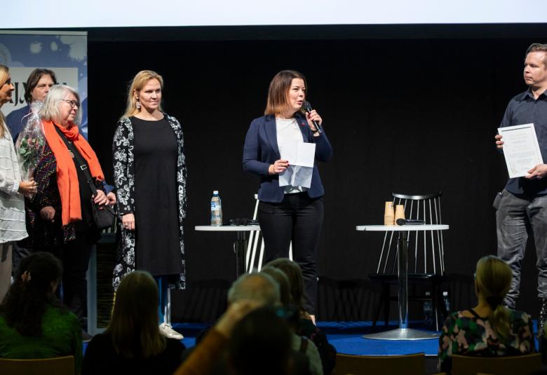 Kuvassa ovat keuruulaiset neljä henkilöä lavalla vasemmalla vastaanottamassa palkintoa. Keskellä on nainen puhumassa mikrofoniin ja oikealla on mies, joka pitää kunniakirjaa kädessään. Alempana on yleisöä selin.