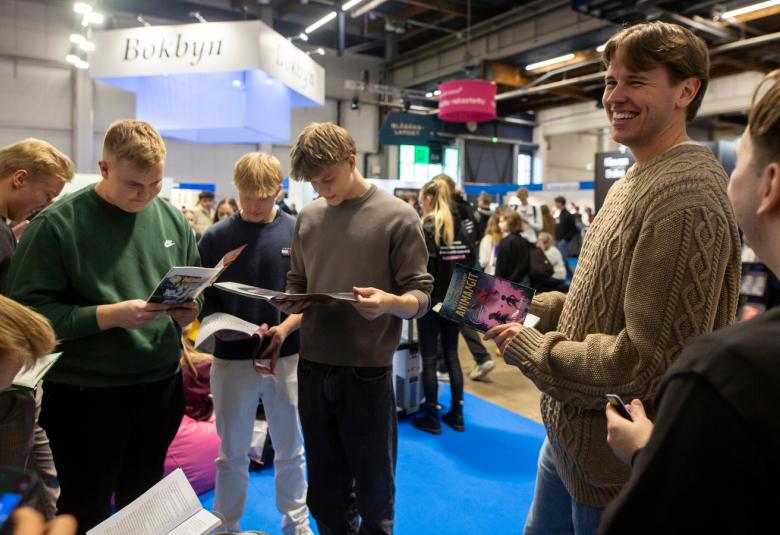 Kuvassa on yläluokkalaisia poikia lukemassa kirjaa. Oikealla näkyy mies, joka katsoo vasemmalle. 