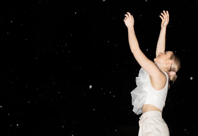 Kuvassa on oikealla Ilta valkoisessa röyhelöpuserossa ja pitkissä housuissa sivuttain kädet ylhäällä. Taustalla on musta tähtitaivas.