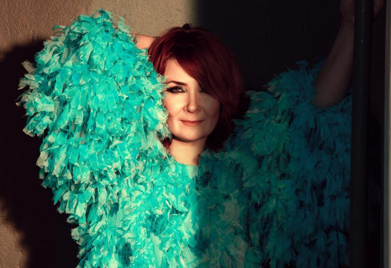 Kuvassa on Irina kädet takaraivossa ja kuva on puolivartalokuva. Hänellä on vihreä pörhöpusero päällään.  Puolet kuvasta on himmennetty.