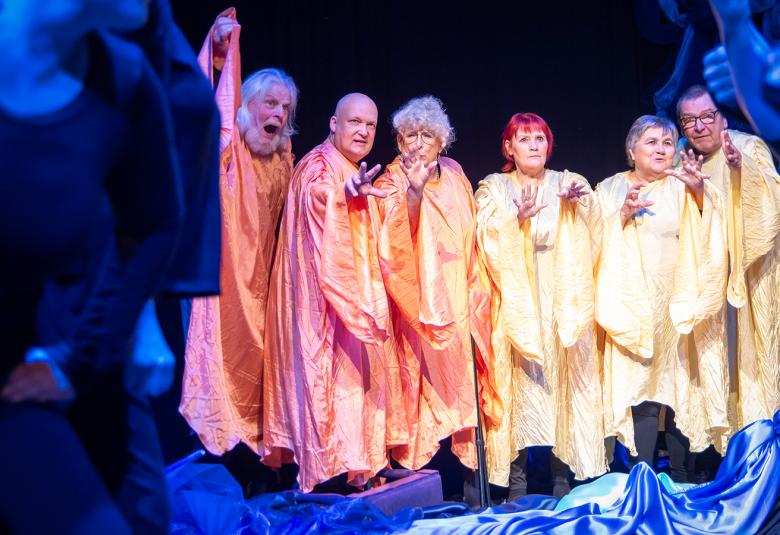 Kuvassa on sinisten kankaiden keskellä vasemmalla kolme punertavissa kauhtanoissa olevaa henkilöä ja oikealla kolme keltaisissa kauhtanoissa seisovaa henkilöä.