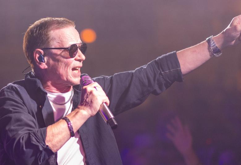 Kuvassa on Kake Randelin laulamassa sivuttain mikki oikeassa kädessään. Vasen käsi osoittaa eteenpäin suorana. Hänellä on musta pusakka, valkoinen t-paita sekä tummat lasit. Tausta on tumma.