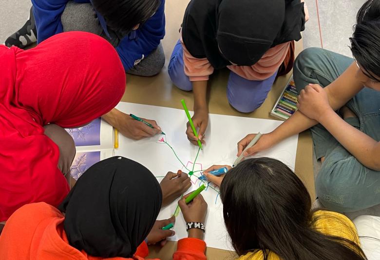 Kuvassa on neljä tummatukkaista nuorta ympyrässä ja heidät on kuvattu ylhäältä päin.  Heillä on yksi paperi ja he piirtävät siihen.