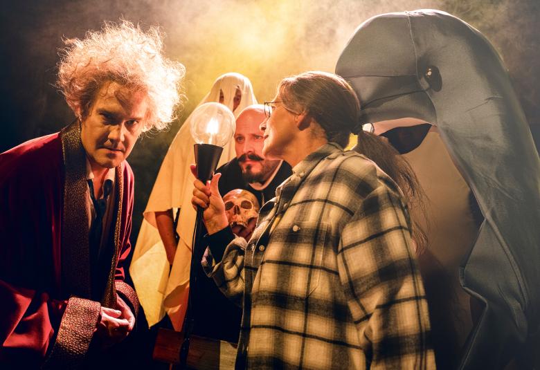 Kuvassa on vasemmalla Samuli Muje violetissa aamutakissa ja hiukset pörrössä. Vieressä on ruutupaidassa lamppu kädessään Teija Auvinen ja takana Hiski Vihertörmä. Takana on valkoinen haamu. Tausta on tumman ruskea ja keskellä on keltaista valoa.