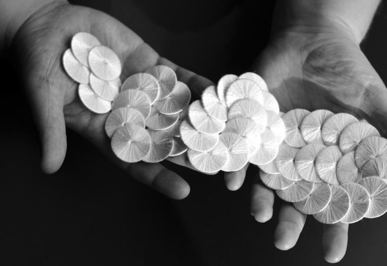 Kuvassa ovat kädet, joiden pääällä on neljässä osassa oleva isokukkainen koru. Keskellä on vaalea kukka. Tausta on musta ja kuva on musta-valkoinen.