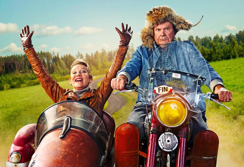 Kuvassa on vasemmalla moottoripyörän sivuvaunussa ruskeassa pusakassa Jaana Saarinen kädet ylhäällä. Oikealla Heikki Kinnunen ajaa moottoripyörää sinisessa pusakassa ja karvahattu päässään. Taustalla on vihreää heinäpeltoa ja metsää.