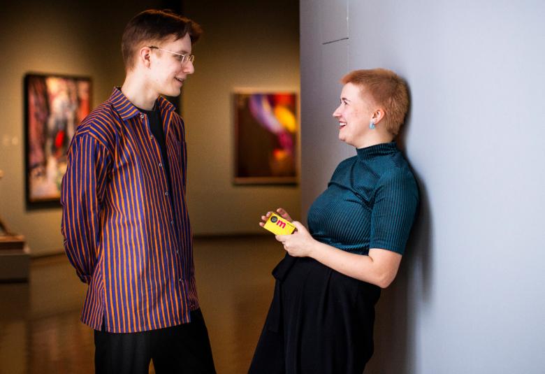 Kuvassa on vasemmalla sivuttain nuori mies tumman puna-siniraidallisessa paidassa seisomassa.  Hän keskustelee edessään olevan naisen kanssa, jolla on tumman sininen paita ja musta puolihame. Hänellä on museokortti kädessään. Taustalla näkyy veistos ja museon tauluja.