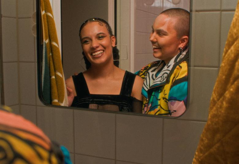 Kuvassa on ruskehtavalla kaakeliseinällä peili, josta näkyy kahden nuoren naisen kasvot. Oikealla näkyy ruskehtavaa pyyheliinaa.