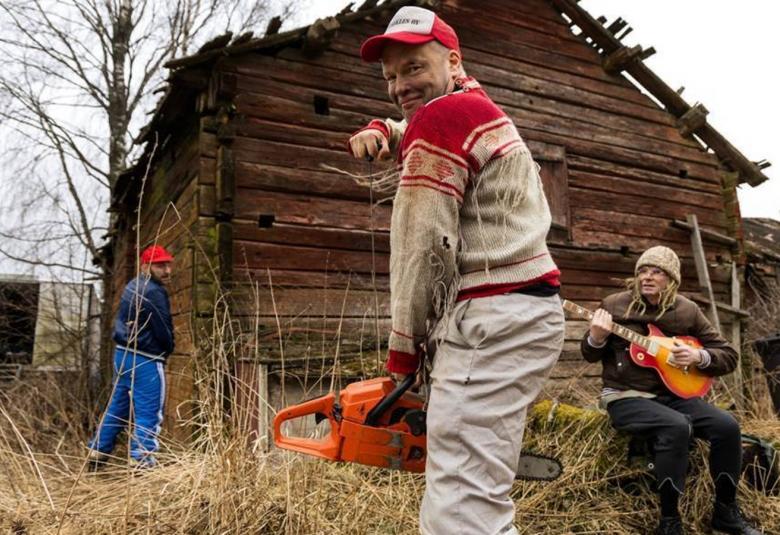 Kuvassa on nuori mies vetämässä moottorisahaa käyntiin ja hänellä on pohjalainen Jussi-paita päällään. Päässä on lippalakki. Taustalla on ruskea huonokuntoinen hirsiaitta ja sen vieressä selin pissalla tumman sinisessä pusakassa ja sinisissä verkkareissa toinen mies. Kannon päällä oikealla on nuori mies vaaleassa pipossa soittamassa kitaraa.