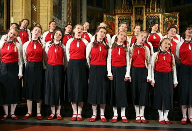Kuvassa ovat Shchedryk-lapsikuoron laulajat kahdessa rivissä. Heillä on punaiset puserot, joissa on valkoiset hihat ja mustat hameet sekä punaiset kengät. Taustalla näkyy kultaisia pylväitä ja kultakehystettyjen ikonien täyttämää alttaria.