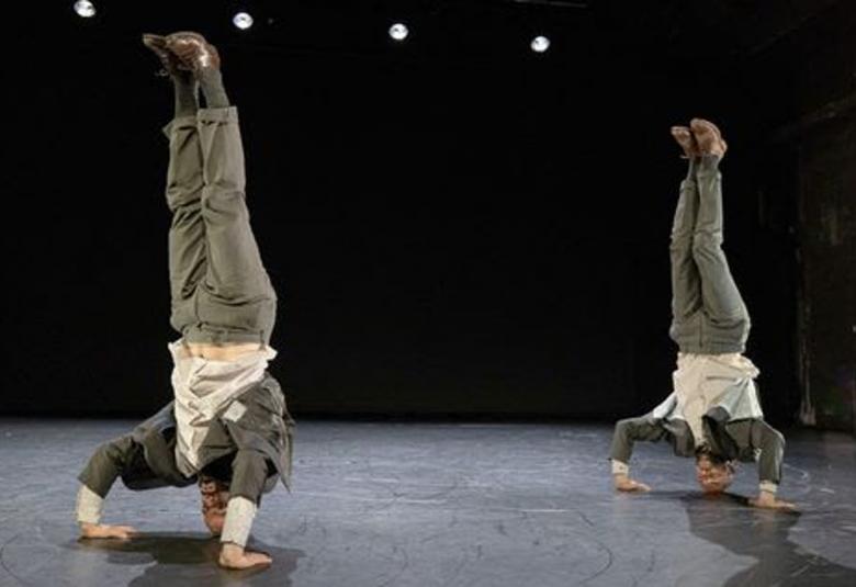 Kuvassa on kaksi miestanssijaa seisomassa päänä päällä ja nojaamassa käsiinsä. Heillä on harmaat housut ja takit sekä vaaleat paidat.  Näyttämön lattia on harmaa ja tausta tumma.