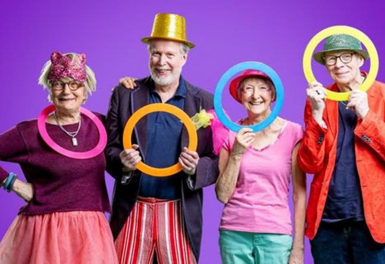 Kuvassa on vasemmalla nainen pinkki rengaskaulassaan ja tumma, burgendin värinen pusero sekä pinkki puolihame. Hänen oikealla puolellaan mies kädessään keltainen rengas , tumma takki ja kultainen silinteri päässään.  Hänen vieressään on nainen, joka katsoo vaalean sinisen renkaan läpi ja pinkissä t-paidassa. Tausta on violetti.