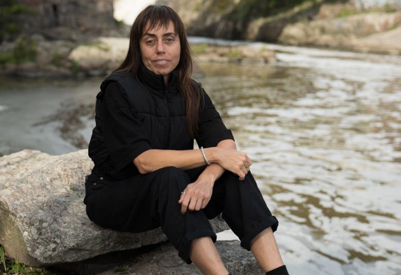 Kuvassa on Zhanna Kadyrova istumassa harmaalla kivellä takaa tulee leveä, harmahtava virta hänen ohitseen. Kadyrovalla on mustat vaatteet ja tumma tukka.