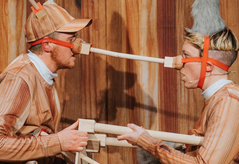 Kuvassa on vastakkain kaksi puuhahmon kaltaista näyttelijää.  Heillä on pitkät puunenät vastakkain toisiaan ja molemmat pitävät kädessään samaa puutuolia. Taustalla on puuseinää.