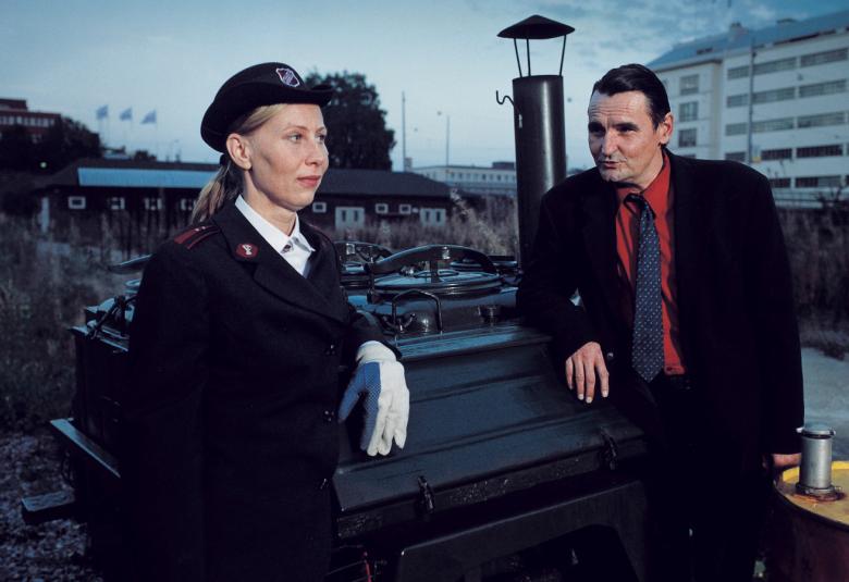 Kuvassa on Kati Outinen vasemmalla tummassa virkapuvussa ja lipallisessa hatussa. Oikealla on mies tummassa puvussa ja tumman punaisessa paidassa. Taustalla on oikealla vaalea kerrostalo ja kaupunkiympäristöä.