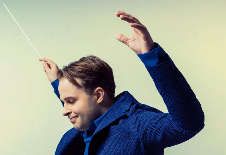 Kuvassa on Taavi Oramo sivuttain johtamassa orkesteria kädet ylhäällä oikeassa kädessään tahtipuikko.  HÄnellä on sininen takki päällään.