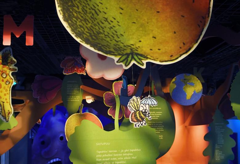 Kuvassa on  vasemmalla kirahvin pää ja yläosassa iso vihertävä omena. Alempana on vihreä iso lehti ja alempana on tumman sinistä. Välissä on mehiläinen. Kuva on värikäs.